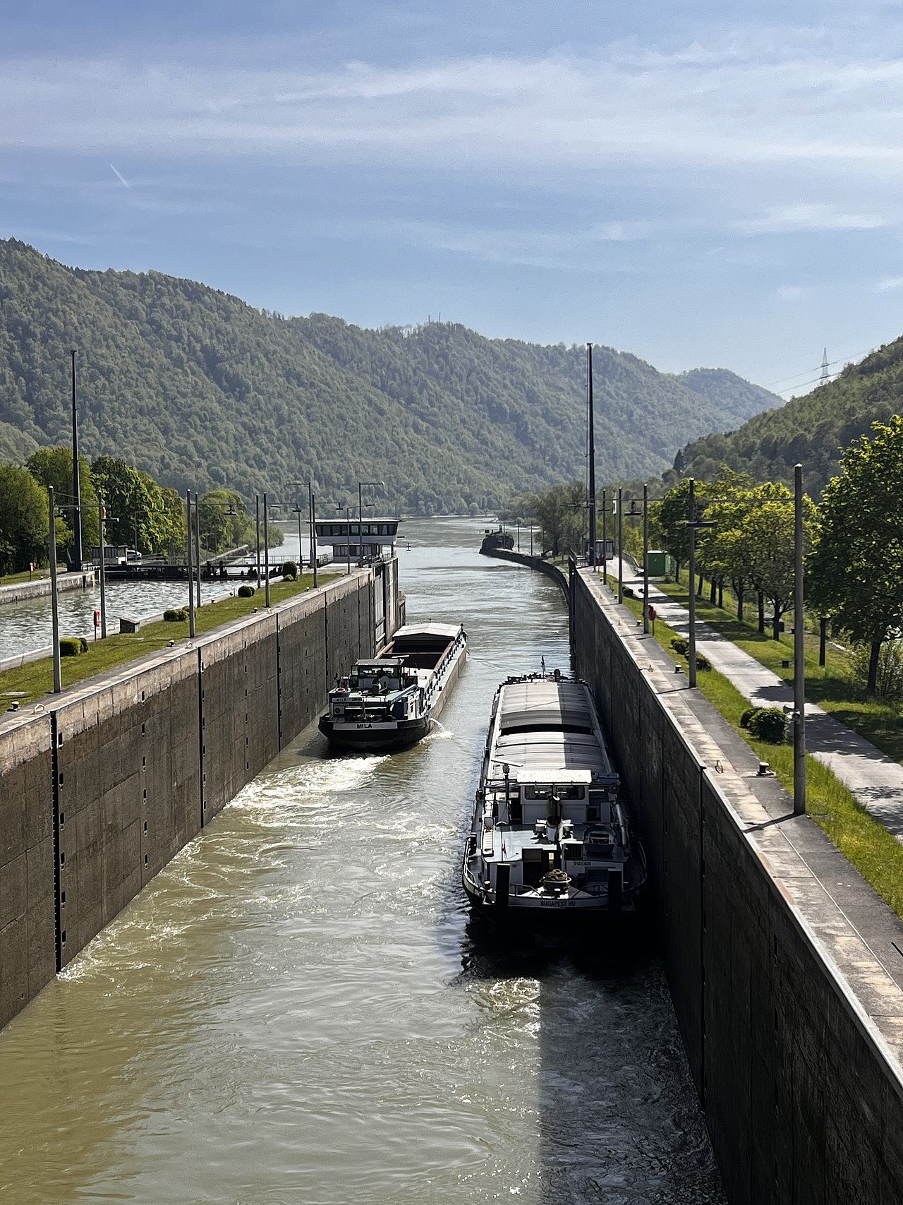 Boats passing through a canal lock in a mountain valley, under a blue sky.