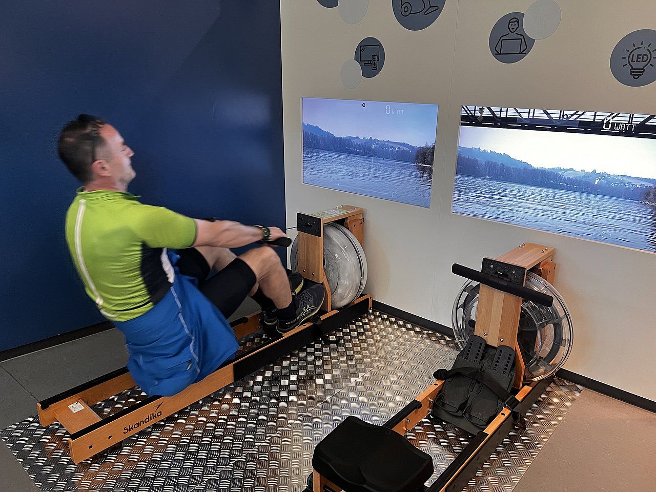 Man rowing on a machine, facing two screens showing snowy landscapes. Indoors, blue wall, silver mat, rowing machines.