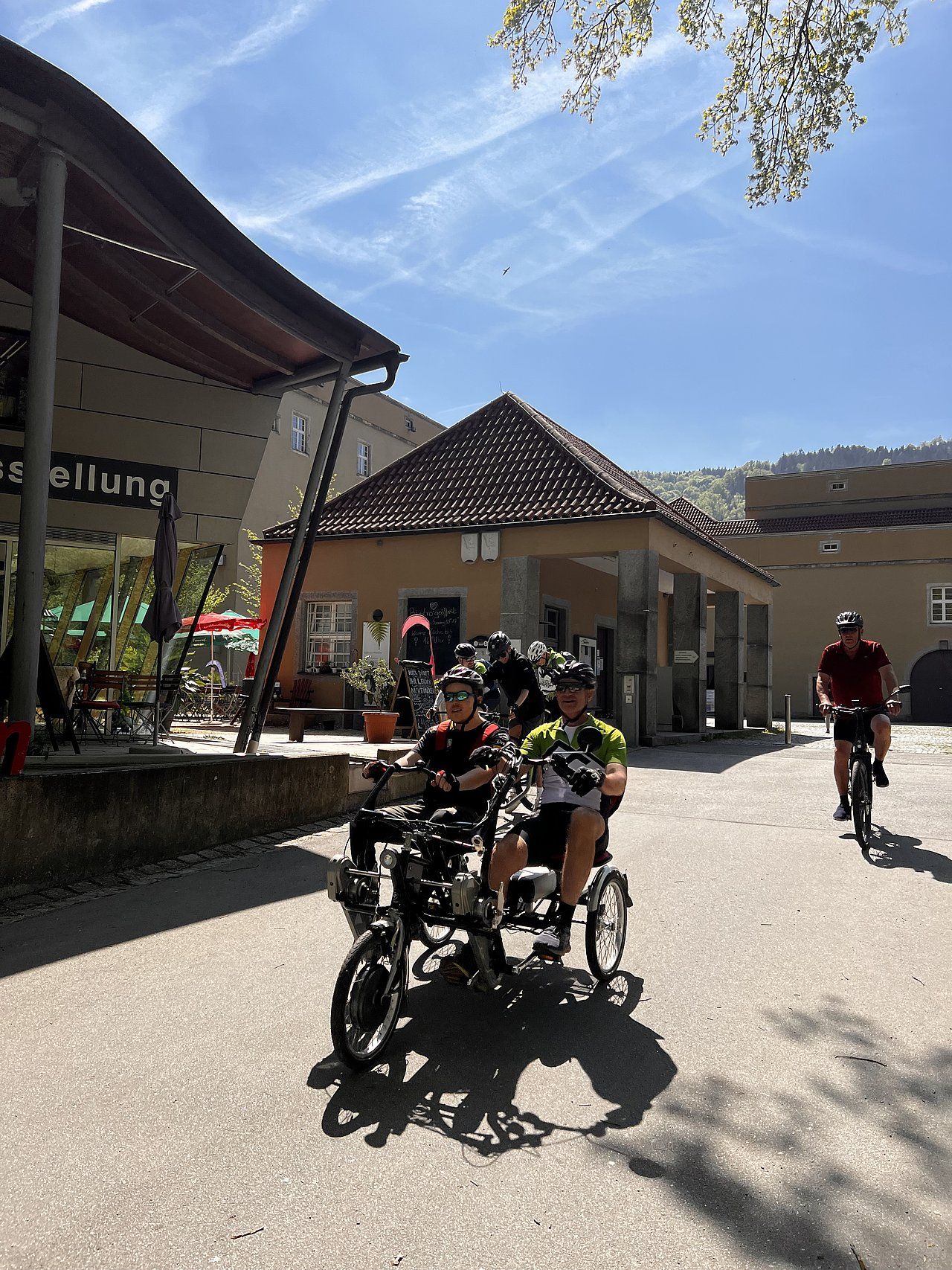 Two people on a tandem bike, one cycling. Buildings, blue sky, and another cyclist in a sunny setting.