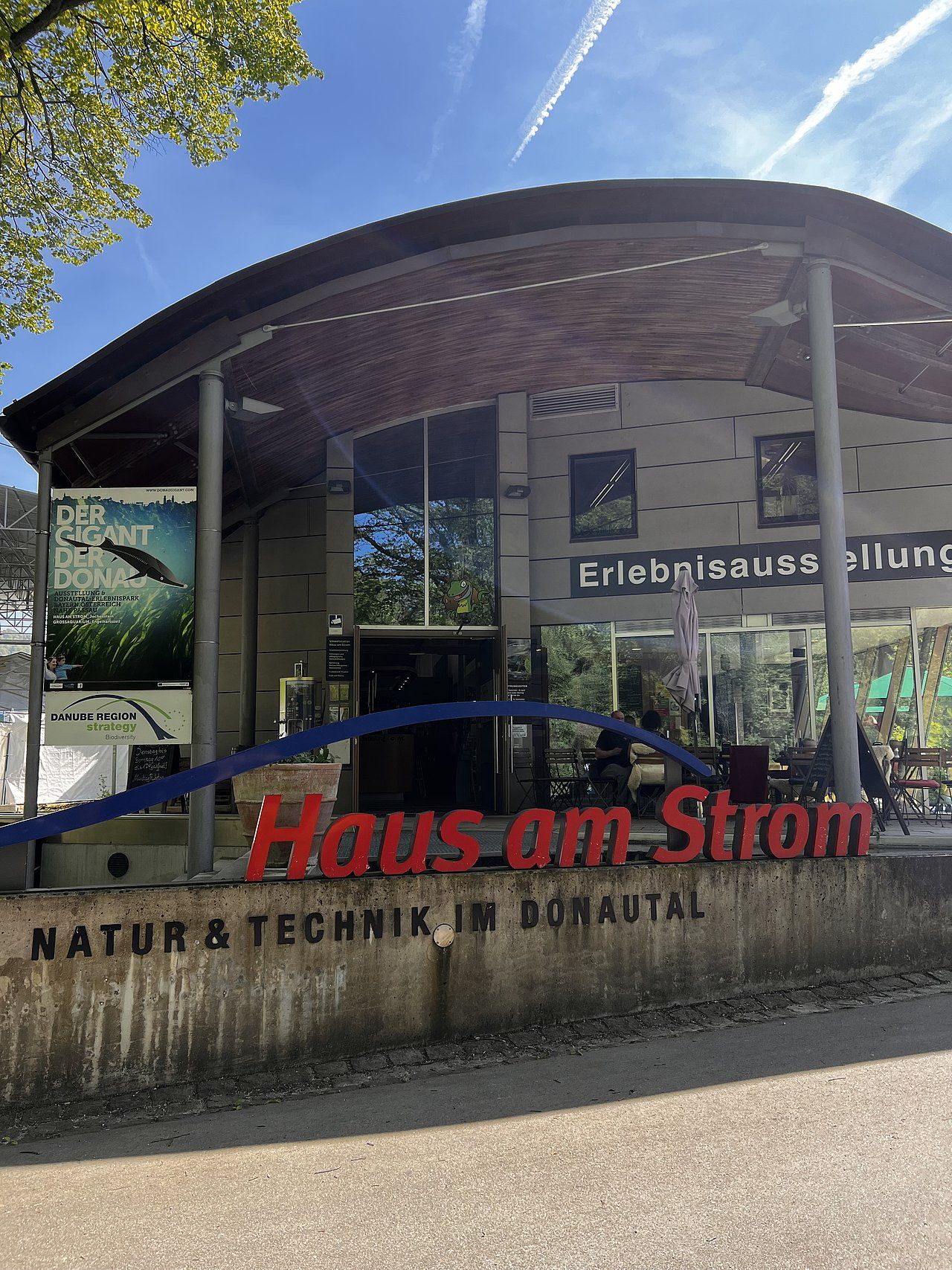 Haus am Strom building with red sign, blue line, and green nature in front.