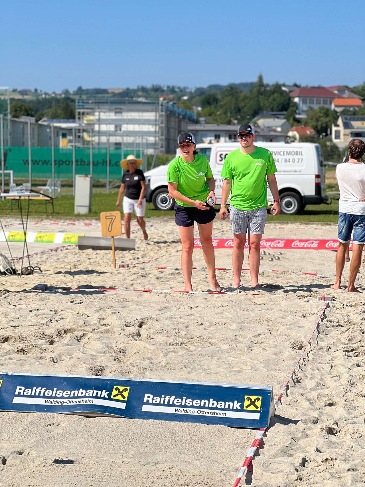 Two people in green shirts on a sandy beach, possibly playing a sport. Other people and buildings in the background.