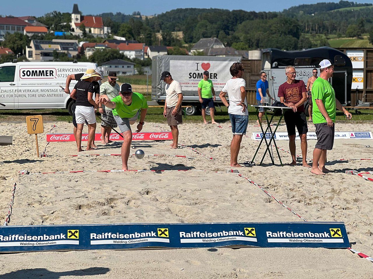 People playing a sand-based game, likely boules or petanque, outdoors on a sunny day.