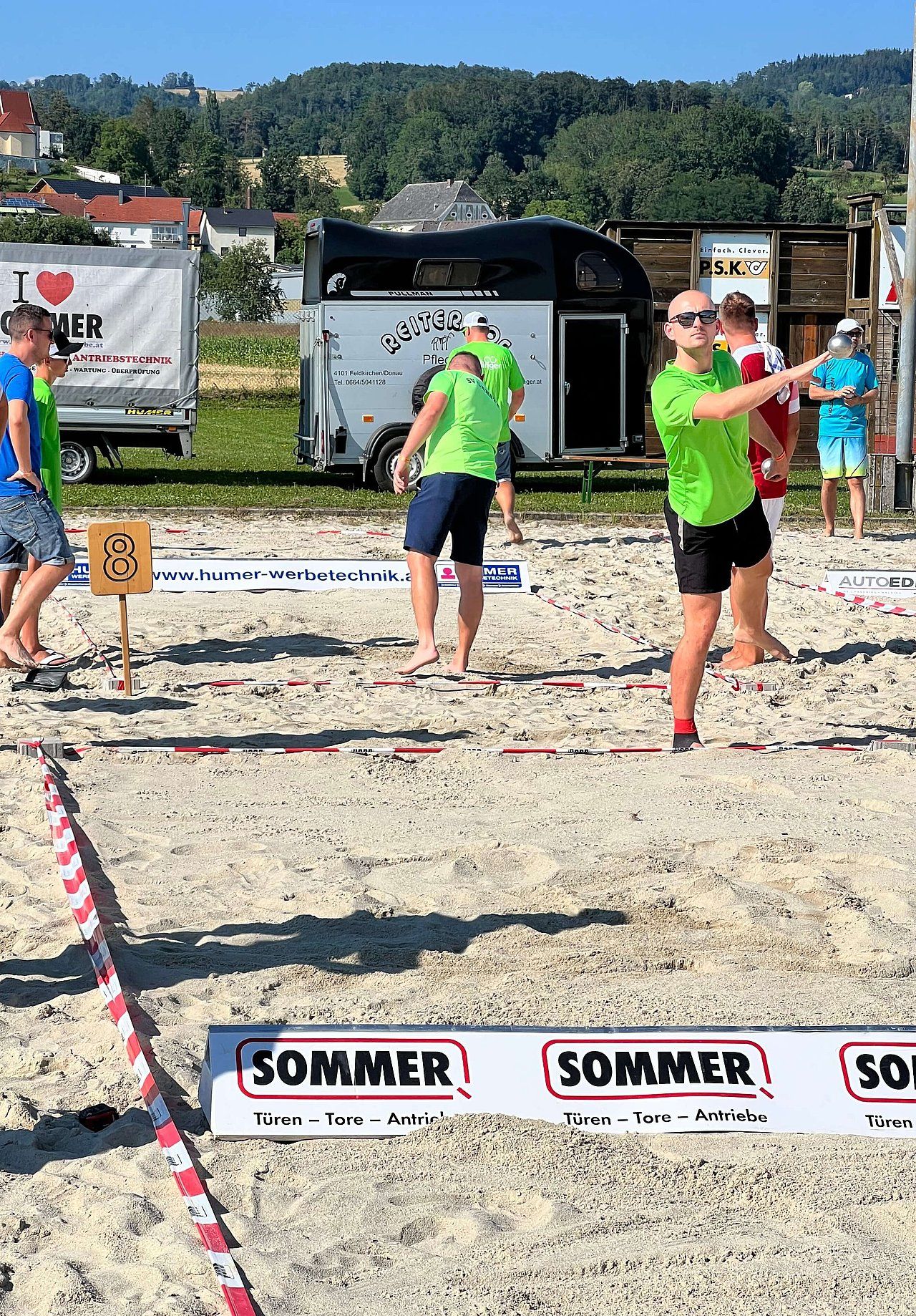 People playing a game on a sandy field. Two in neon green shirts, one throwing, near a trailer and Sommer banner.