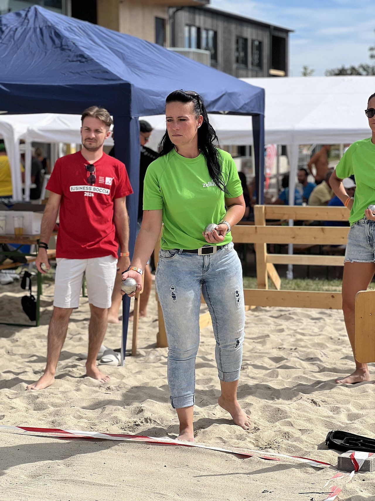 Woman in green shirt throws a metal ball during a game on sandy ground; man in red shirt watches.