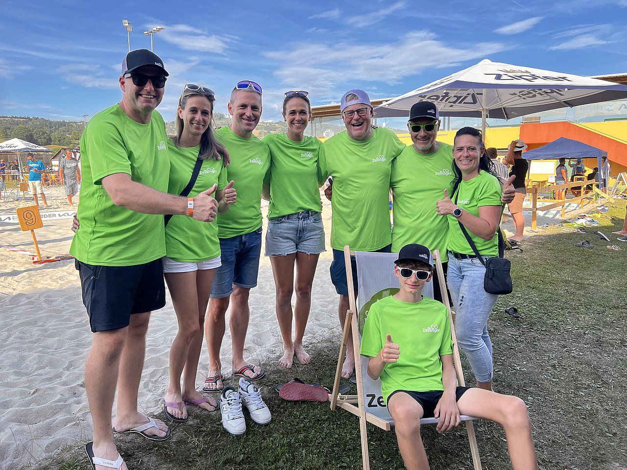 Group of people in green shirts at an outdoor event. Smiling, some giving thumbs up. Beach setting.