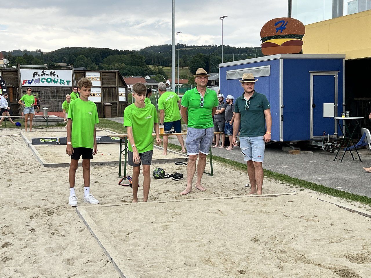 People playing a sand-based game outdoors; two boys in green shirts, two men in green and blue shirts, a burger sign.