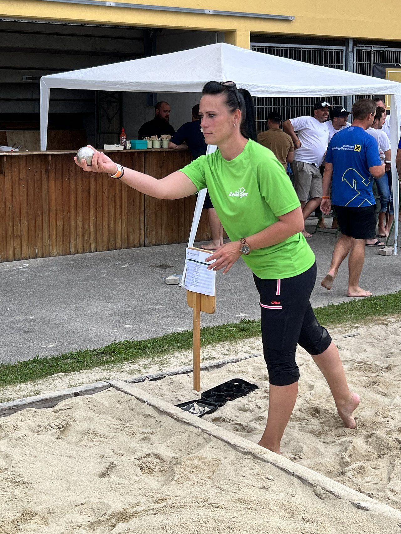 Woman in green shirt throws a metal ball in a sand pit during a game of pétanque.