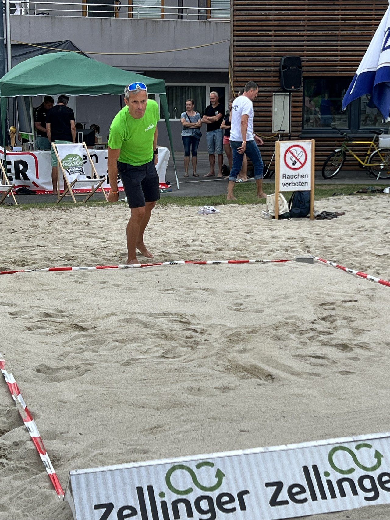 Man in green shirt and shorts playing a game on a sandy court, other people watching.