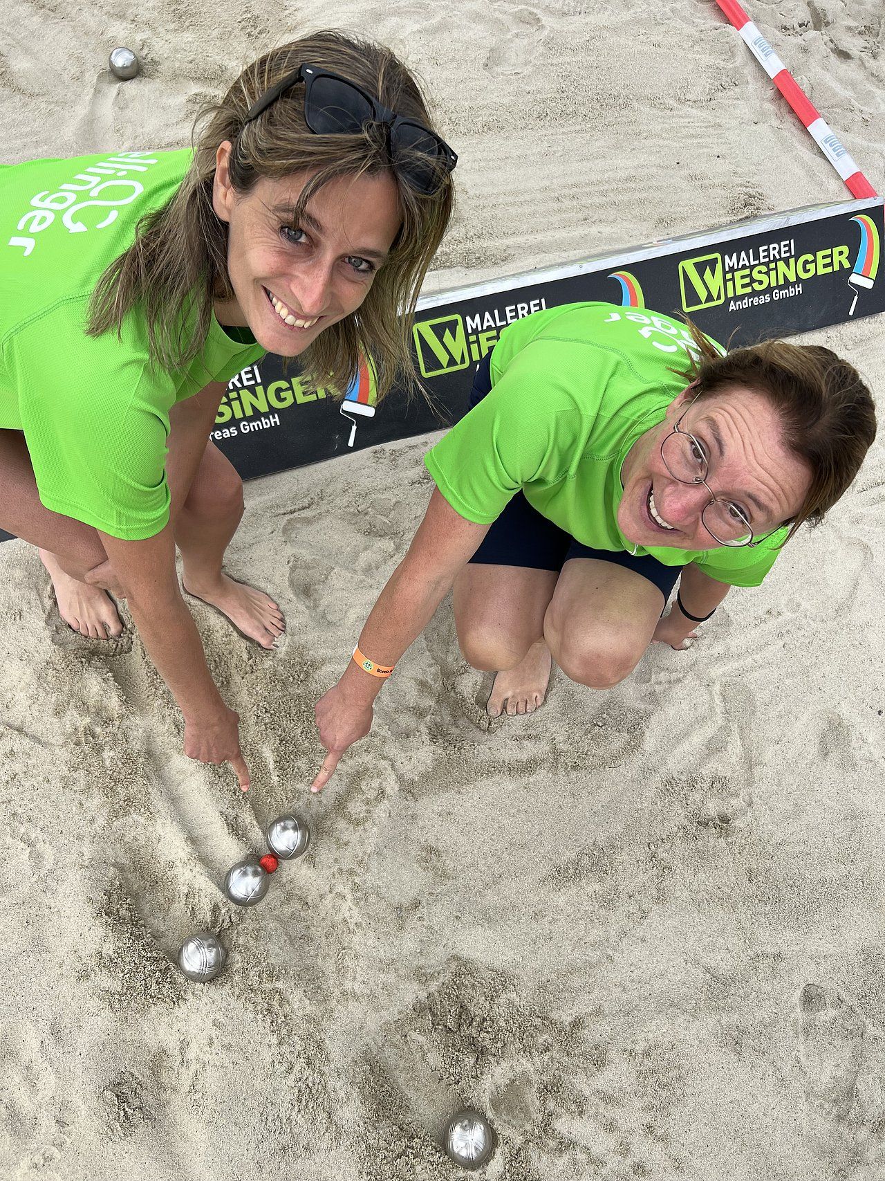 Two women in green shirts playing boules on sand; one points.
