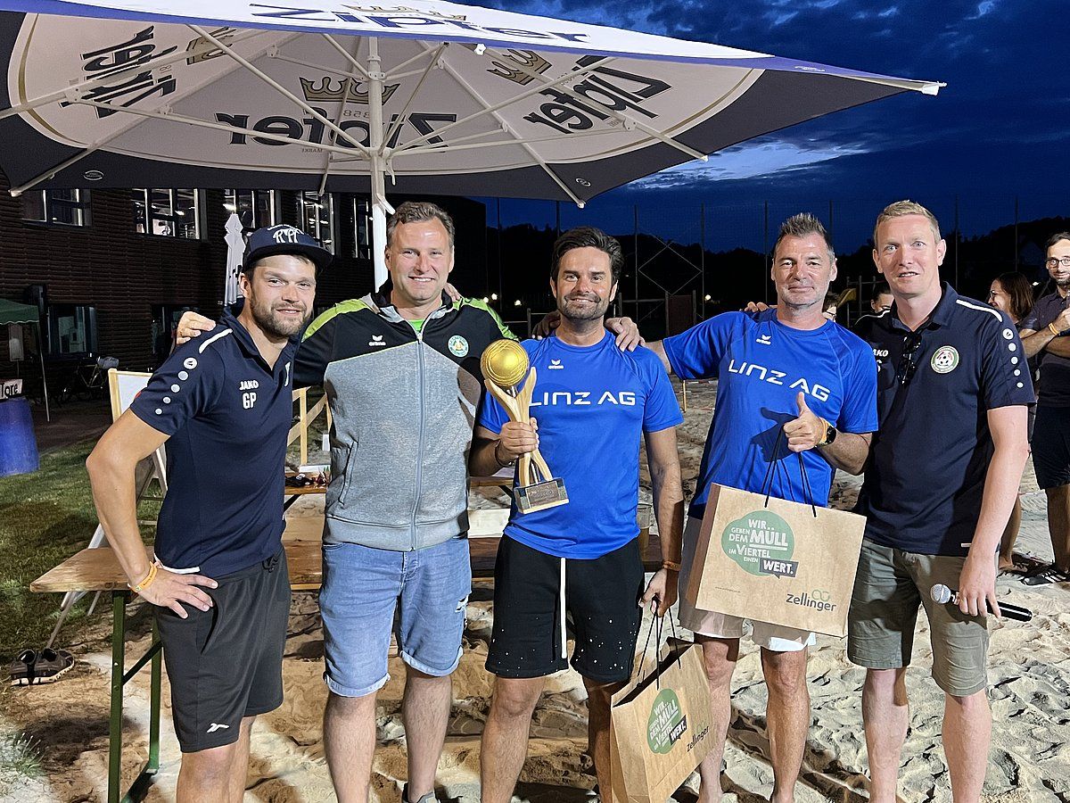 Five men posing outdoors, holding a trophy and bags. Some wear team shirts. Smiles abound.