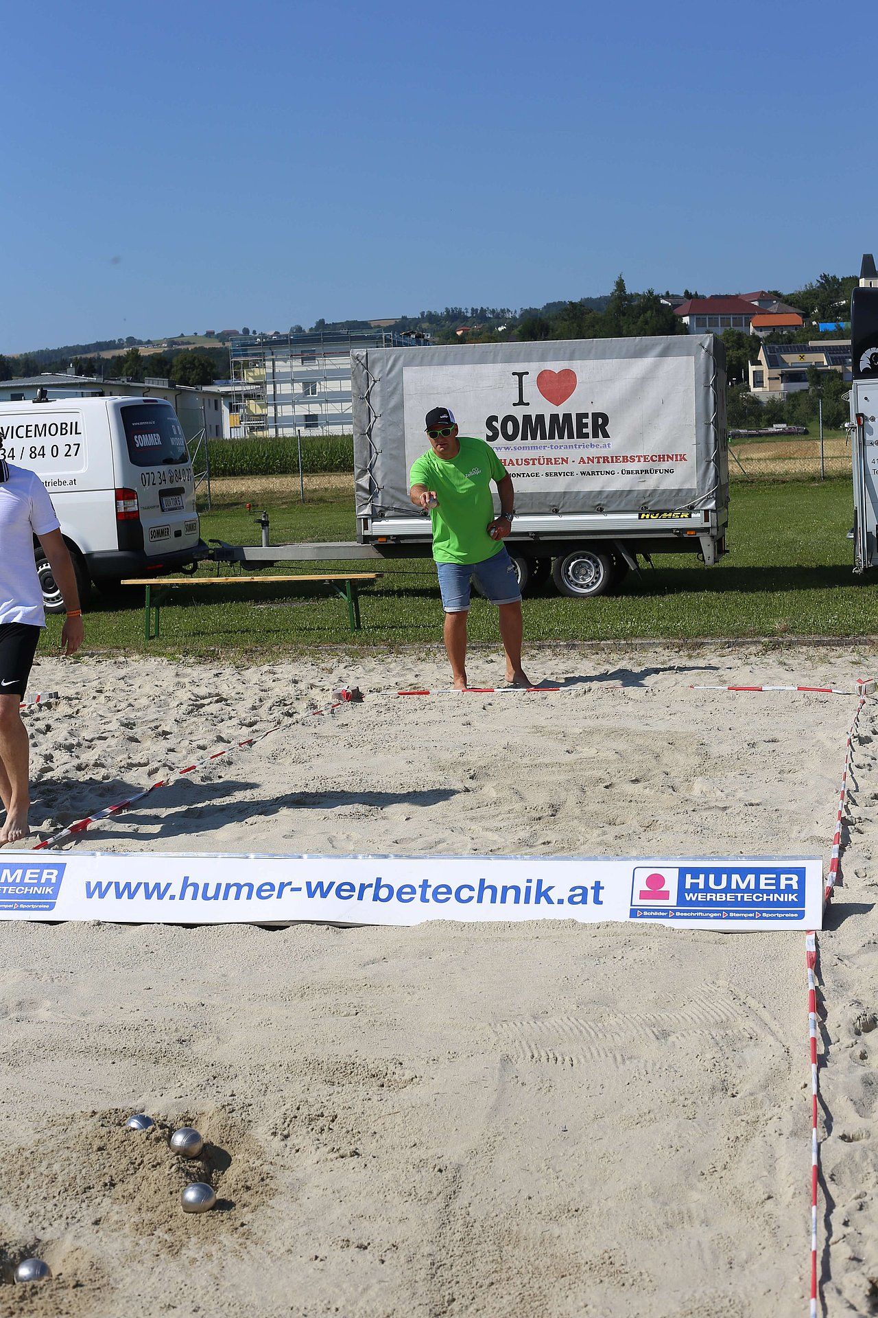 Man in green shirt throws a metal ball in a sand pit; a game of pétanque on a sunny day.