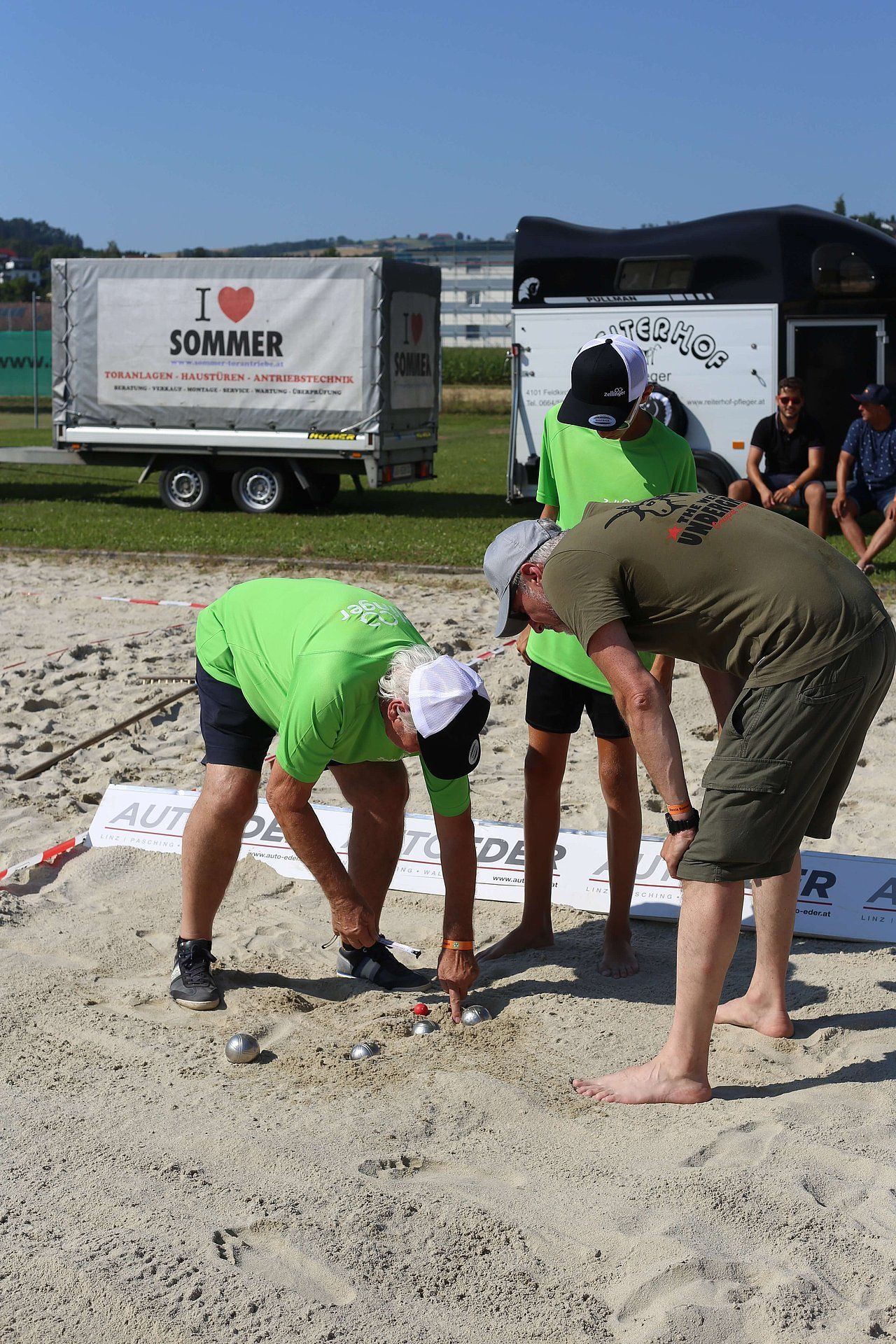 People playing pétanque on a sandy field. One man in green shirt bends to measure. Others watch.
