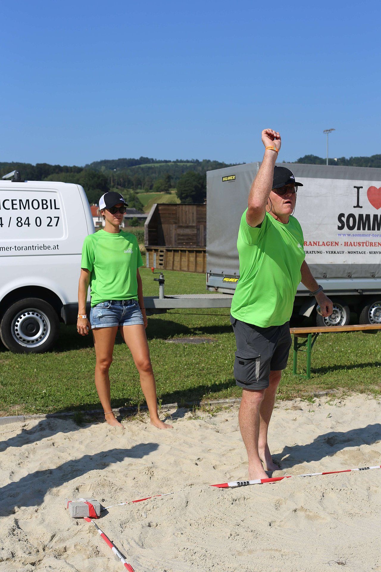 Two people in green shirts playing a game on a sand court, one raising their arm in celebration.