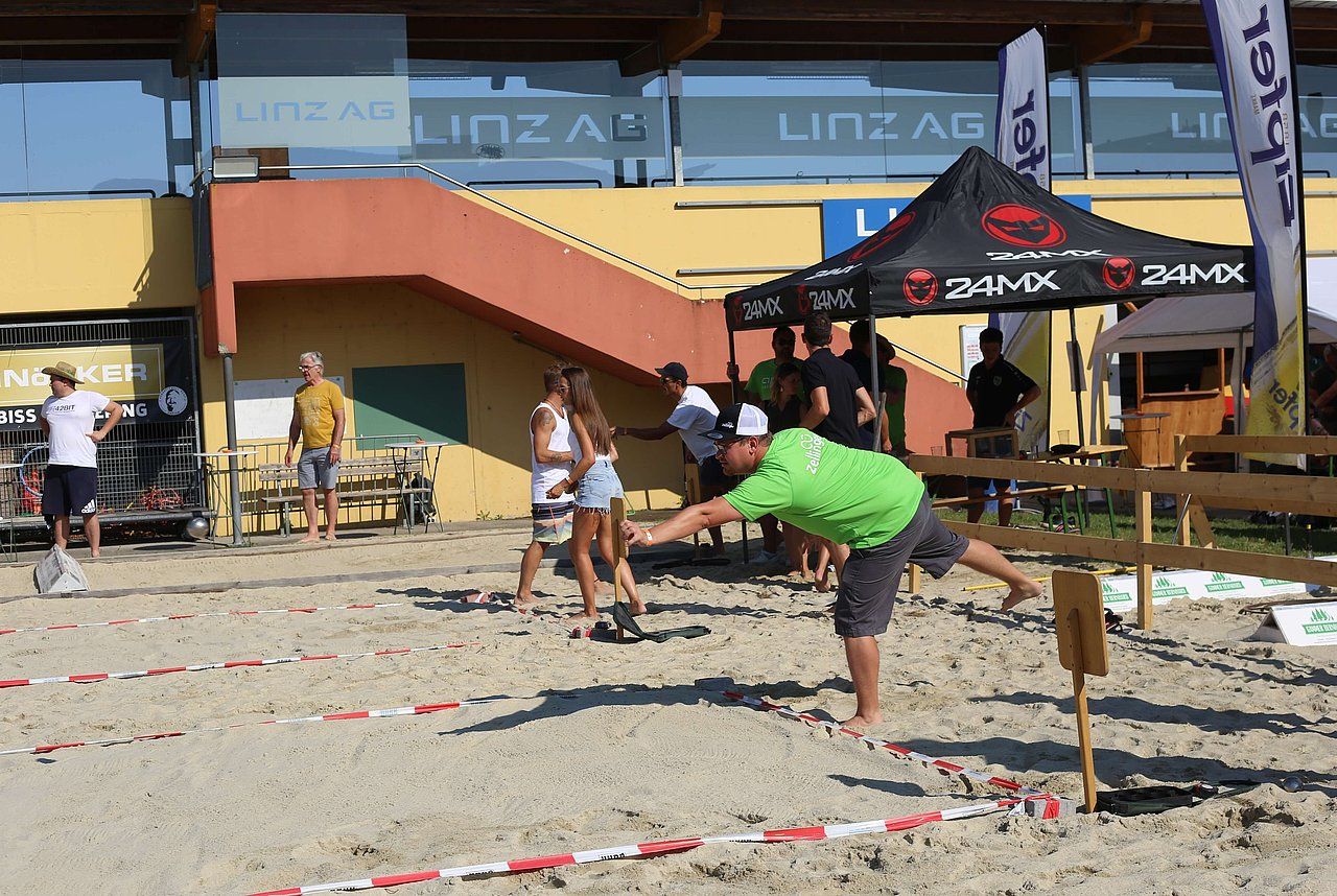 Man in green shirt throwing object on sand at an outdoor event. People watch near tents, under a blue sky.
