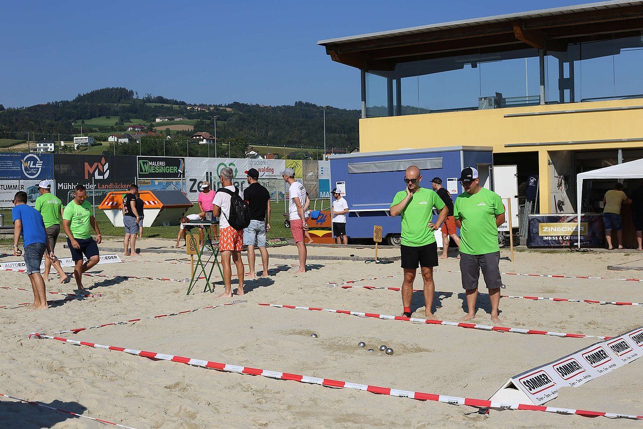 Beach volleyball court with people. Some wearing green shirts, others in shorts. Sunny outdoor setting.