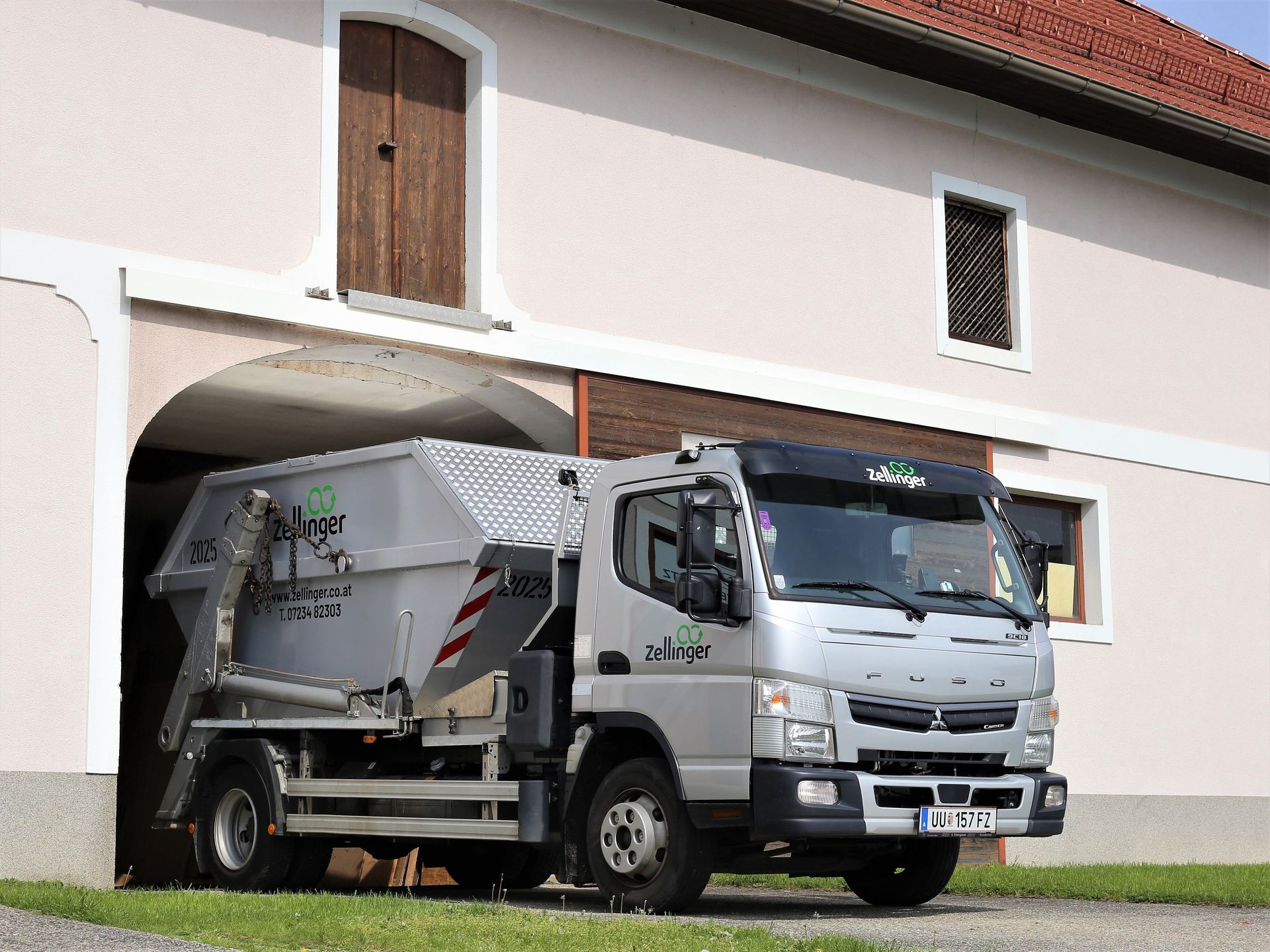 Garbage truck exiting a building archway; silver truck with bin, white building.