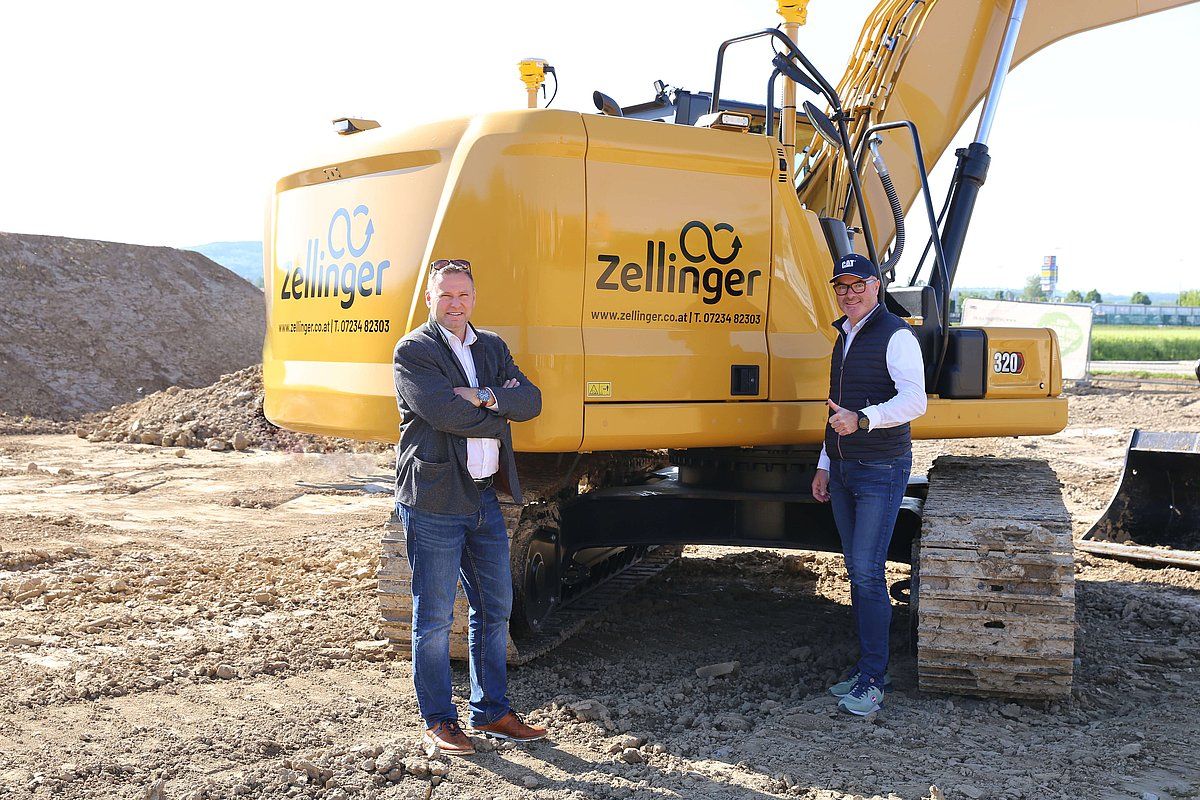 Two men standing in front of a yellow excavator at a construction site.