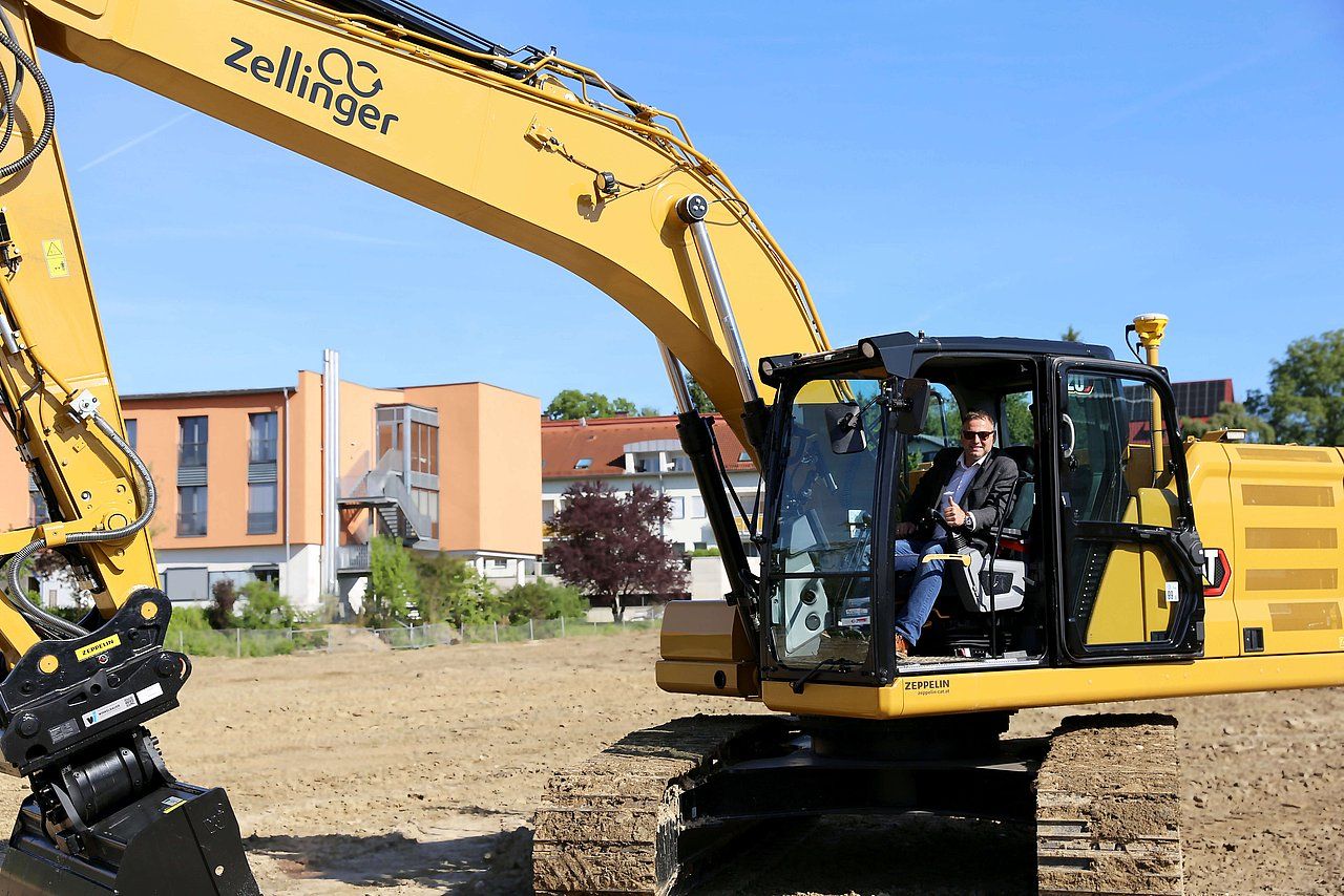 Man in excavator cab on construction site, yellow machine, buildings in background.