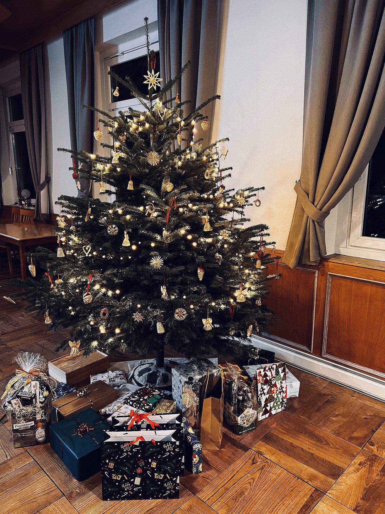 Christmas tree decorated with lights and ornaments, surrounded by wrapped gifts on wooden floor.
