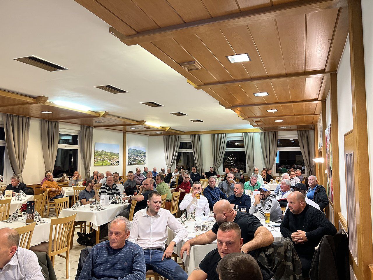 A crowded dining room with people seated at tables; some are talking. Wooden paneling and warm lighting.