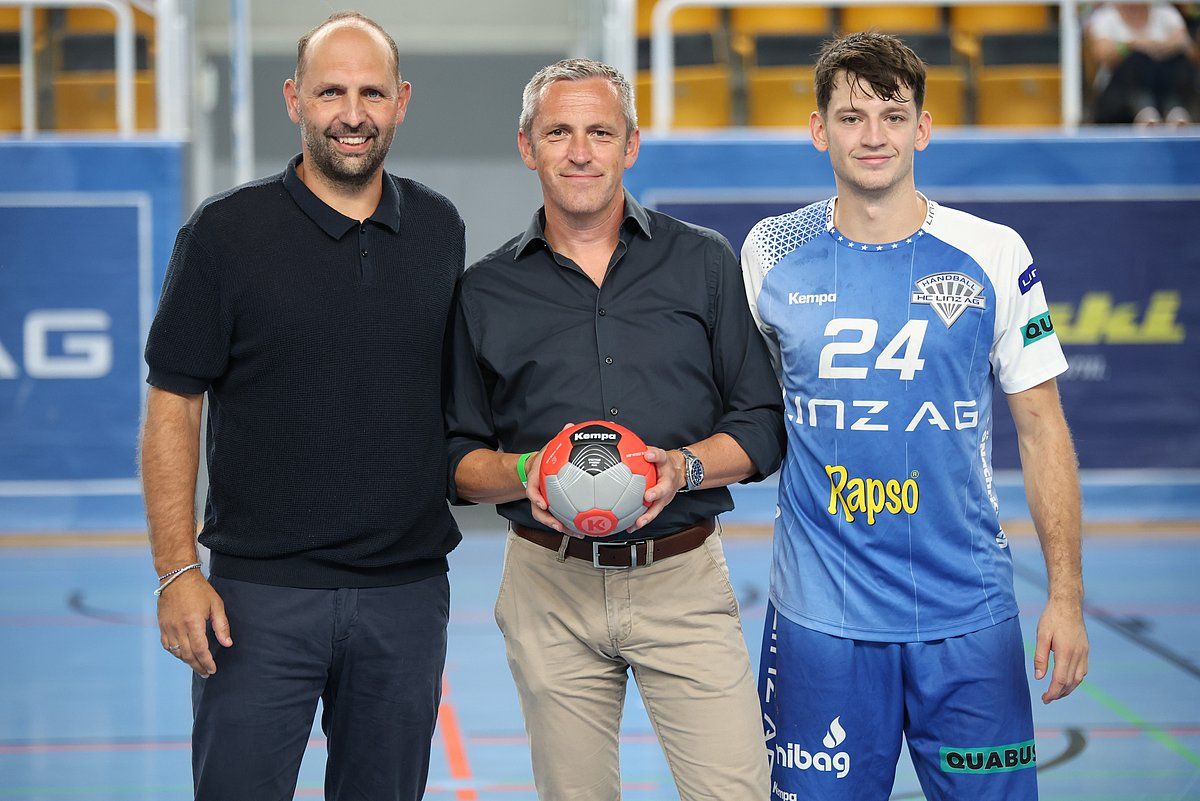 Three men, one holding a handball, pose in an indoor court. The player wears a team uniform.