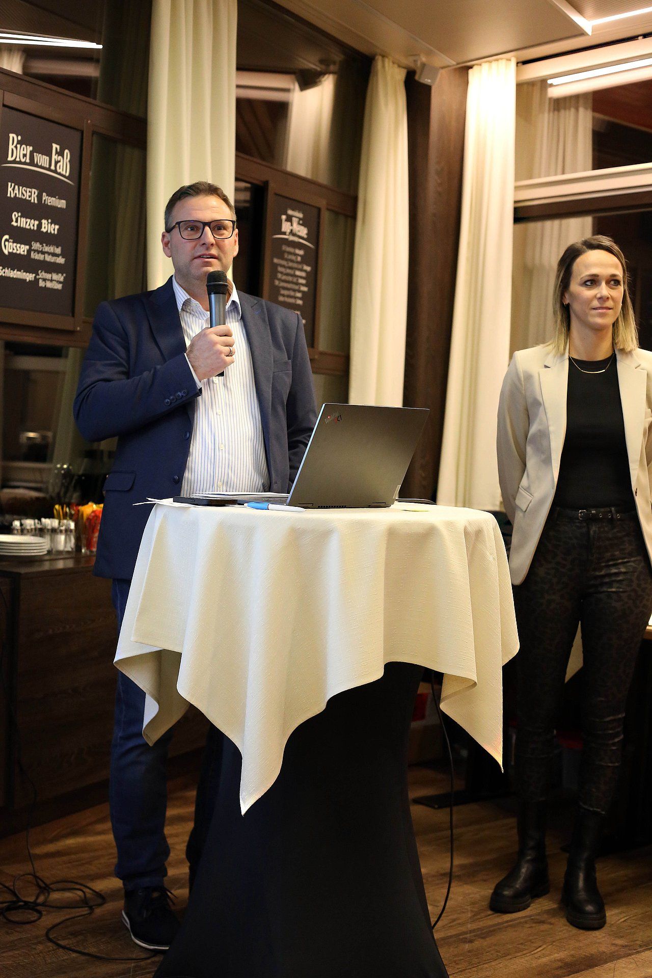 Man speaking at a podium with laptop. Woman stands beside. Indoors, restaurant setting.