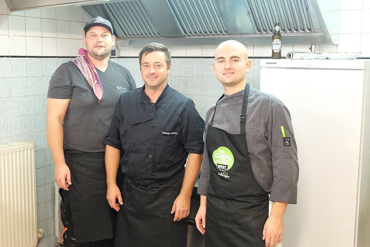 Three chefs in a kitchen, wearing aprons and looking at the camera.
