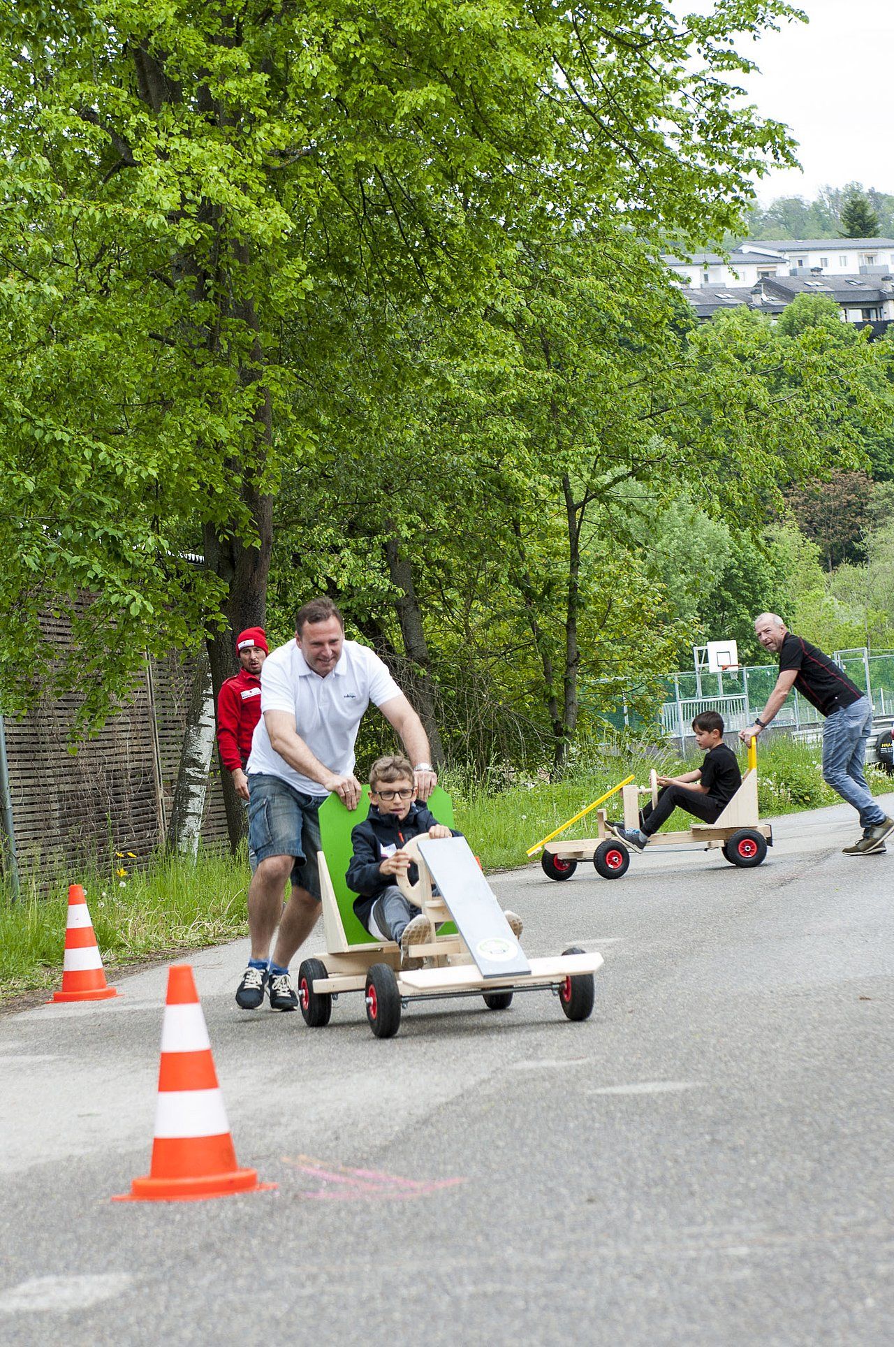 People pushing kids in homemade wooden race cars down a street, with trees and cones.