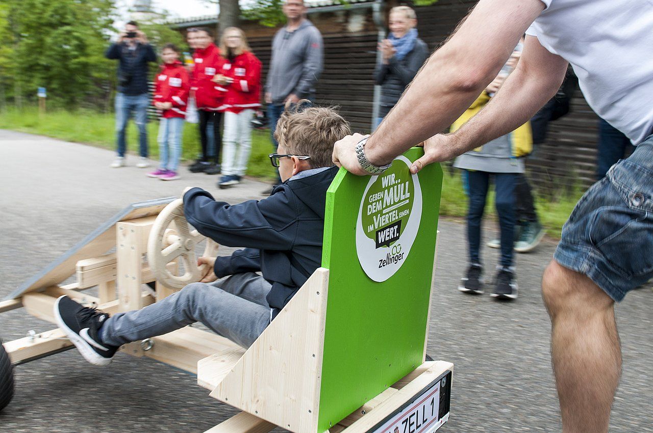 Boy in wooden soapbox car, pushed by an adult, racing on a street, spectators watch.