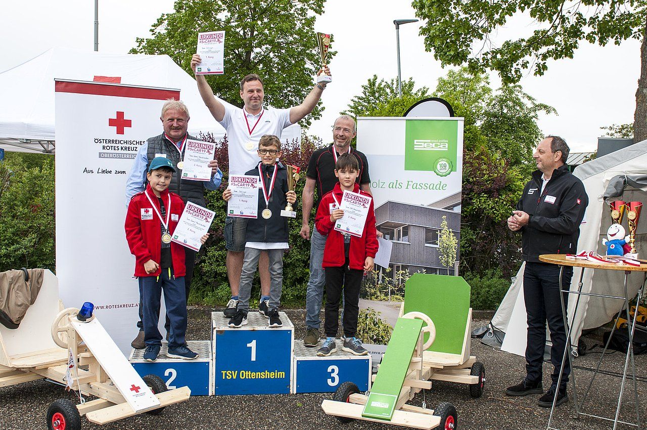 Winners on a podium holding awards, wooden carts nearby, outdoors.