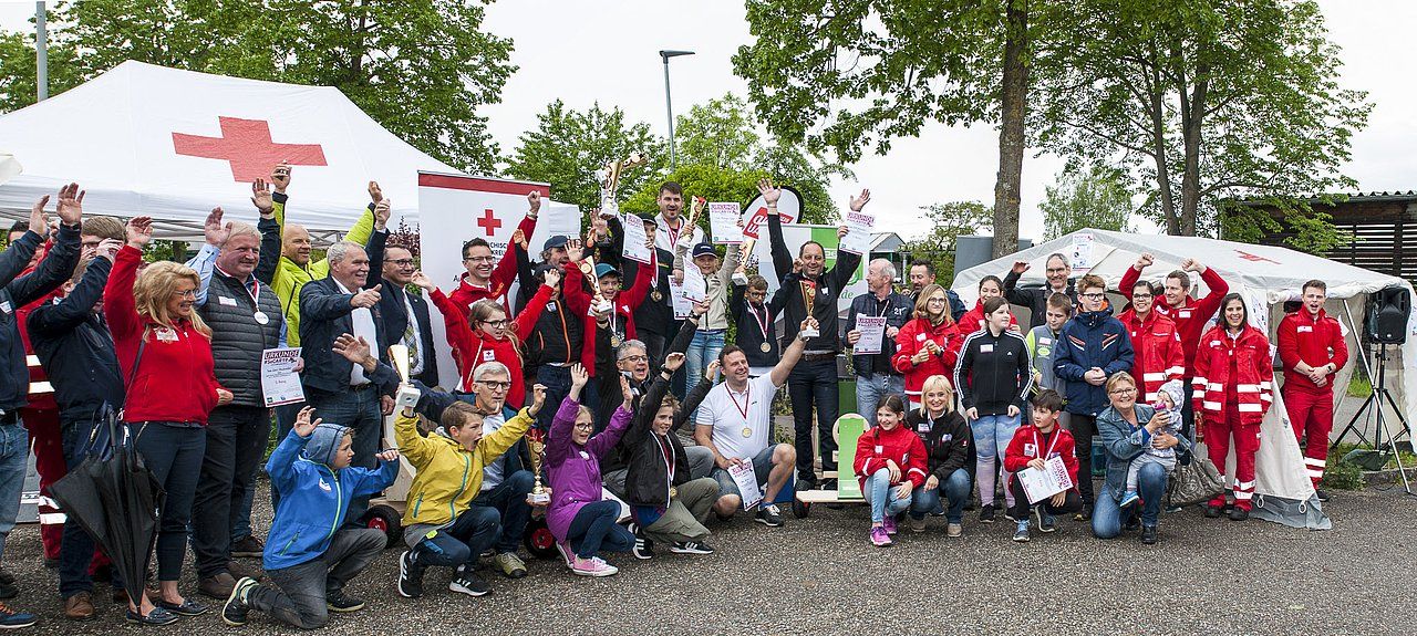 Large group of people, many in red, cheering in front of a Red Cross tent.