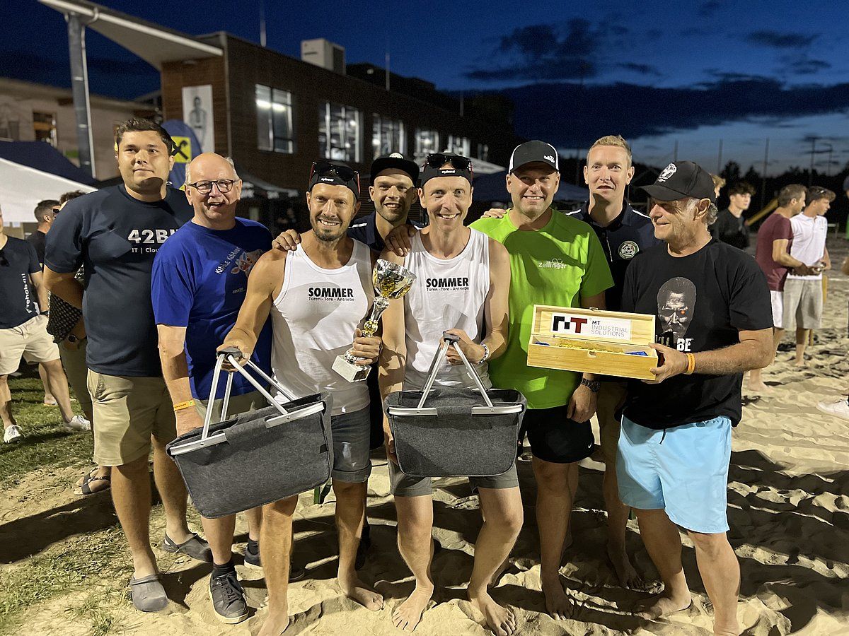Group of men smiling, holding prizes and baskets on a sandy beach, possibly after a competition.