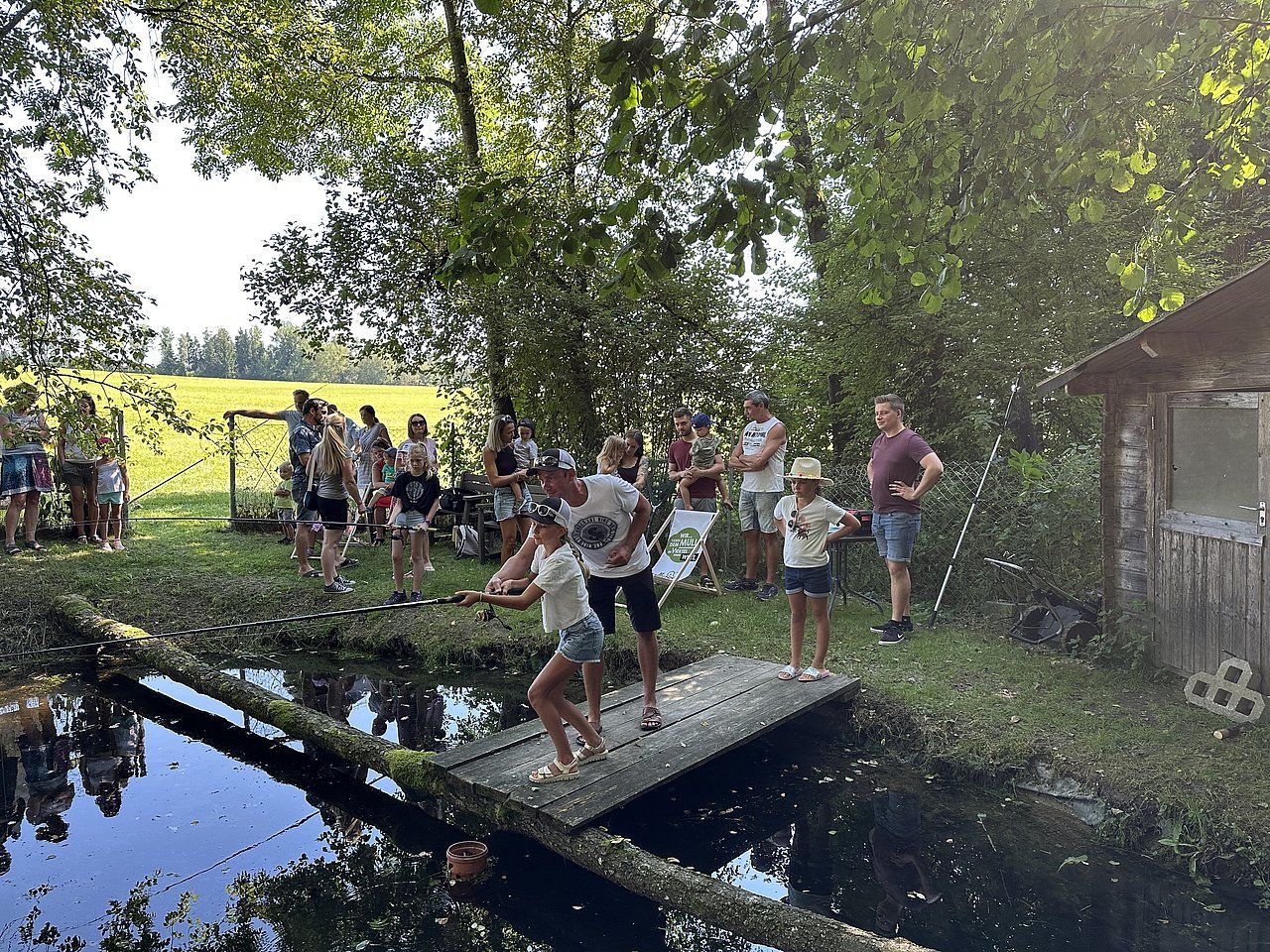 People on a makeshift bridge over water, near a small building. Green trees and field in background.