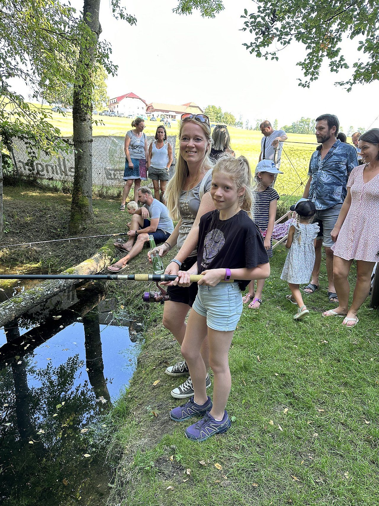 Girl fishing with woman near a small stream, surrounded by onlookers on a grassy bank.