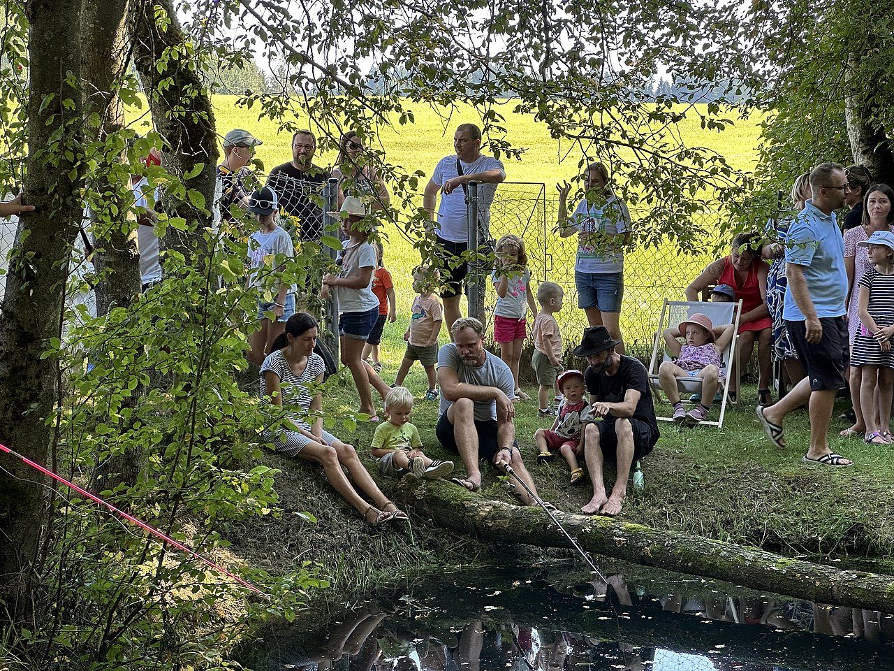 People gathered by water's edge, some sitting, others standing, enjoying a sunny day.