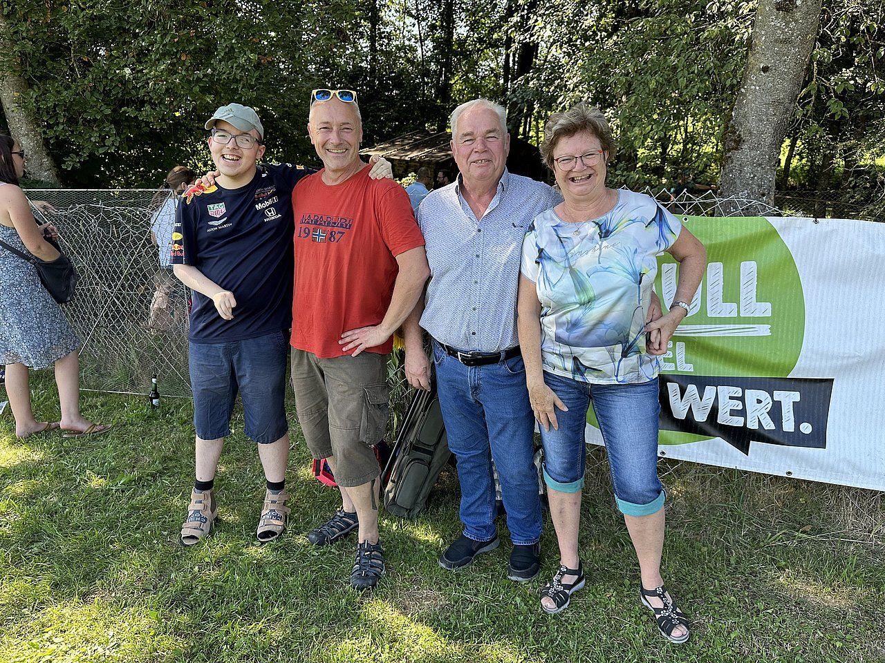 Four people pose outdoors on grass near a sign. They smile, and trees are visible.