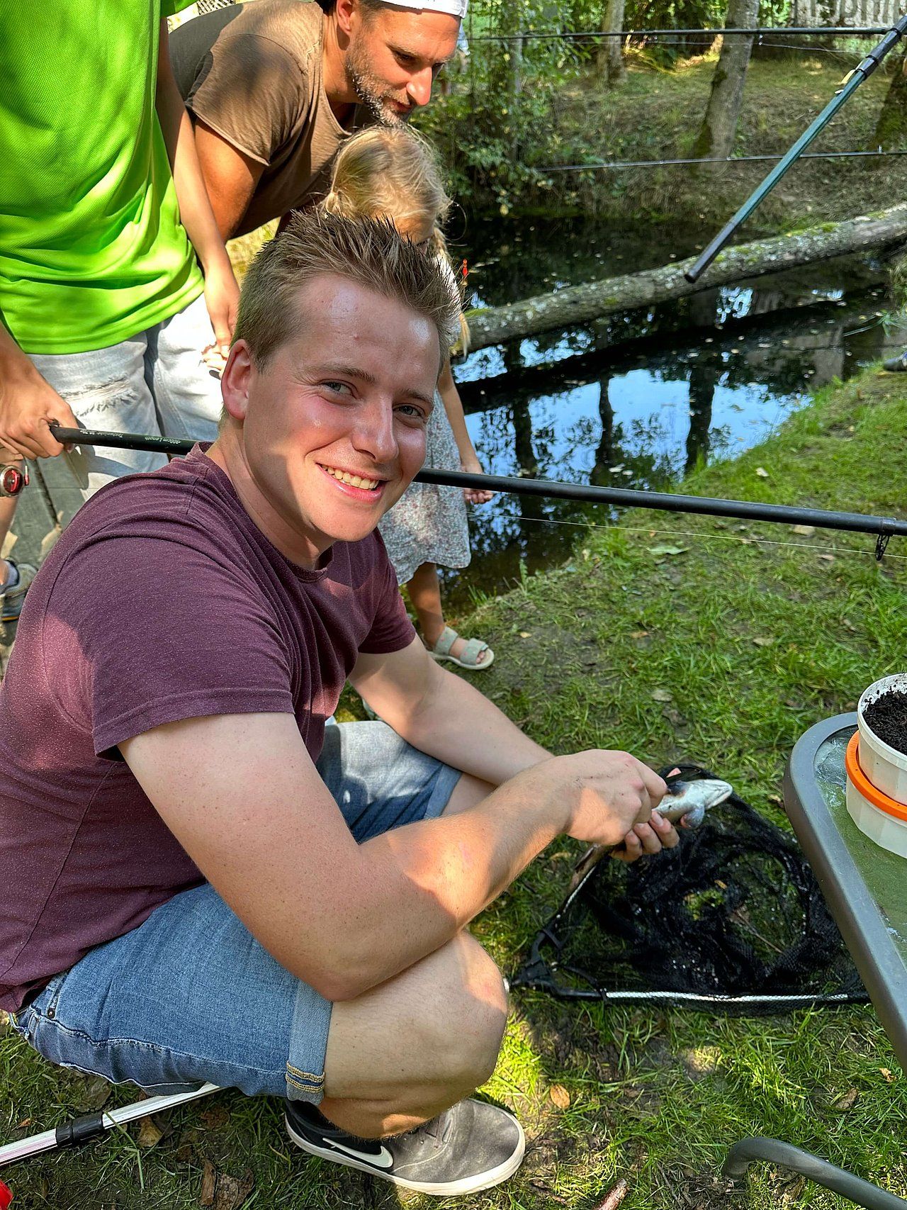 Man kneeling, holding fish in a net, smiling. Other people fishing near a stream, outdoors on grass.