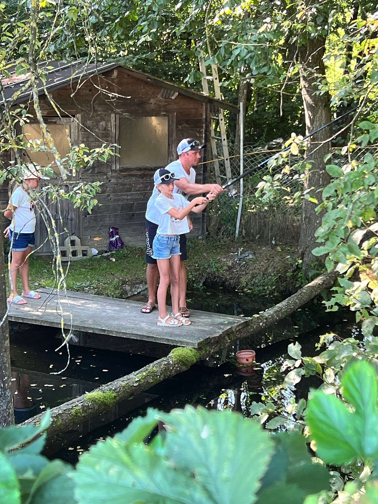 Man teaching a girl to fish from a wooden dock next to a pond. A second girl watches. Outdoors, sunny.