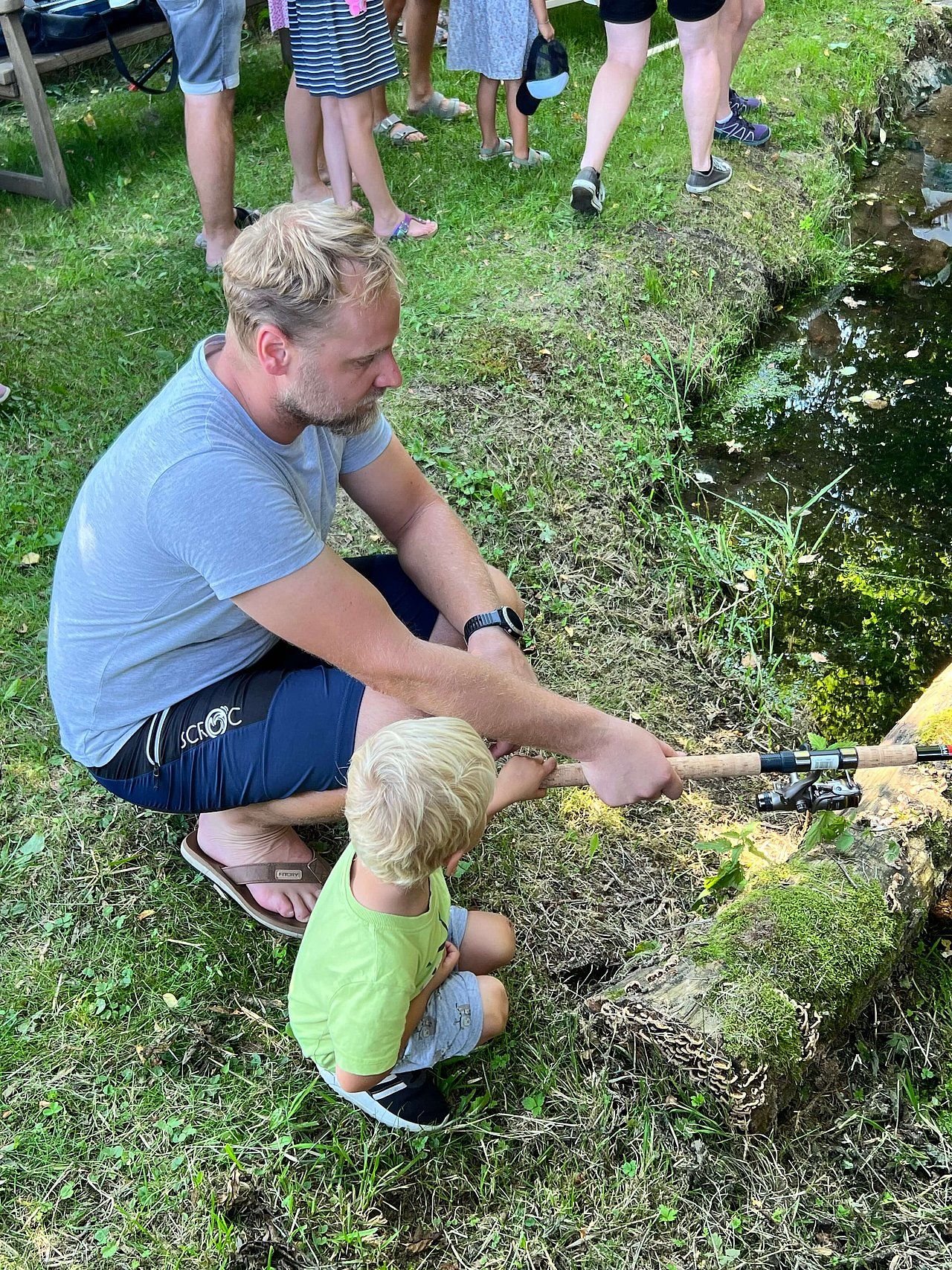 A man and child fish by a pond. The man is kneeling and holding a fishing rod, the child watches.