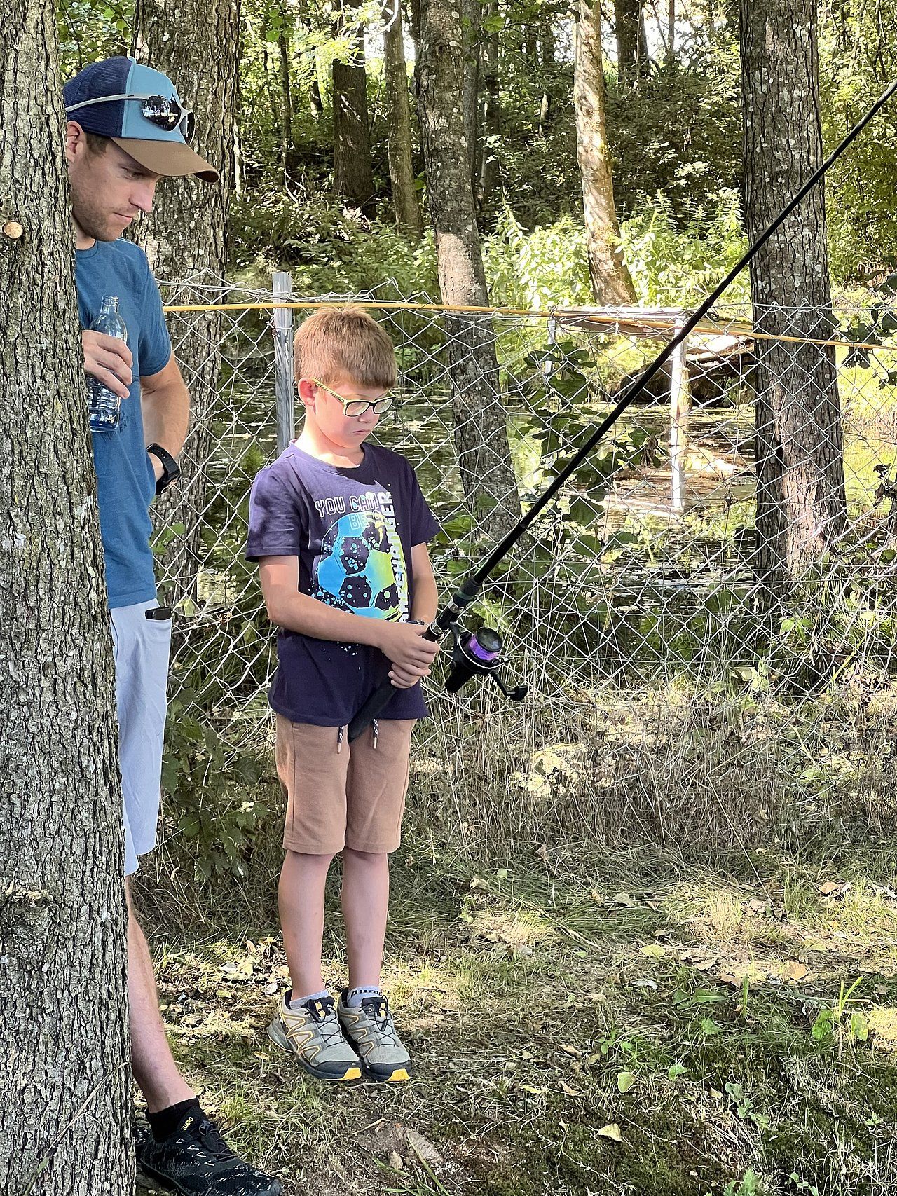 Two people fishing near a chain-link fence. A boy with glasses holds a fishing rod, and a man looks on.