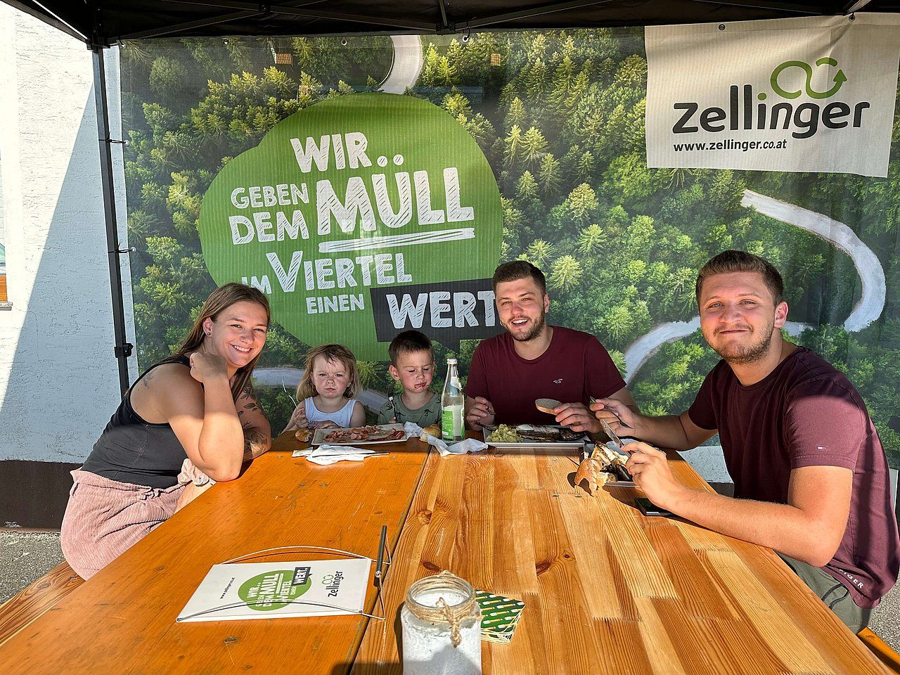 Family eating at a wooden table outdoors, with a banner that says