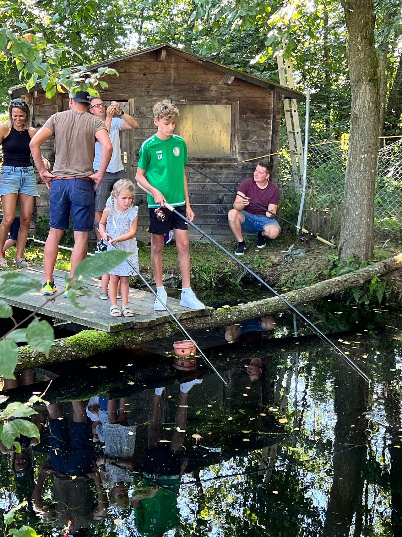 People fishing by a pond near a small wooden shack. One child holds a rod while others watch.