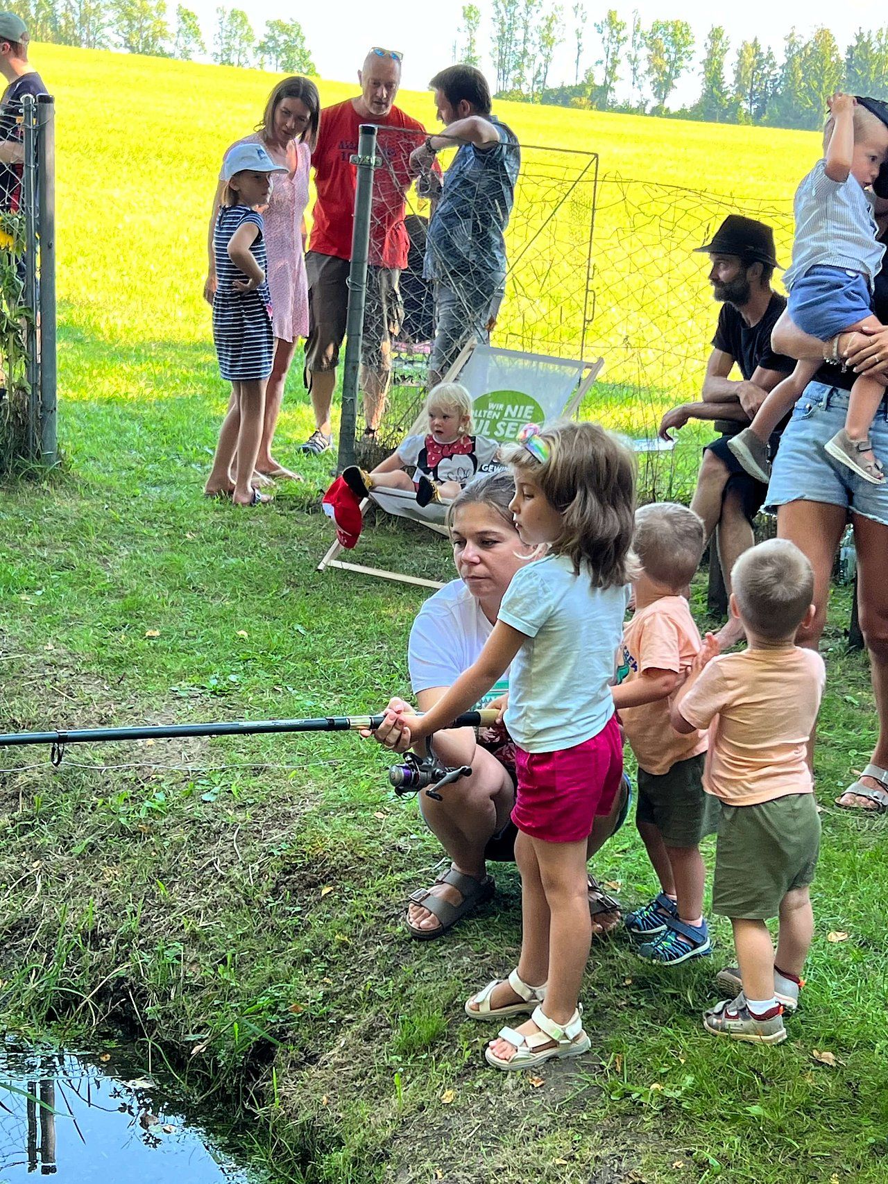 Woman and children fishing; group of people at a grassy outdoor area; sunny day.