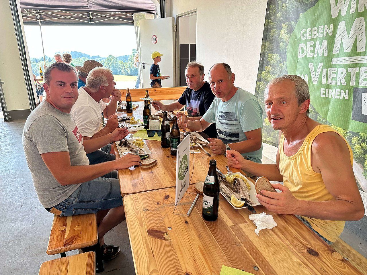 Men eating at a long wooden table outdoors, beers, food.