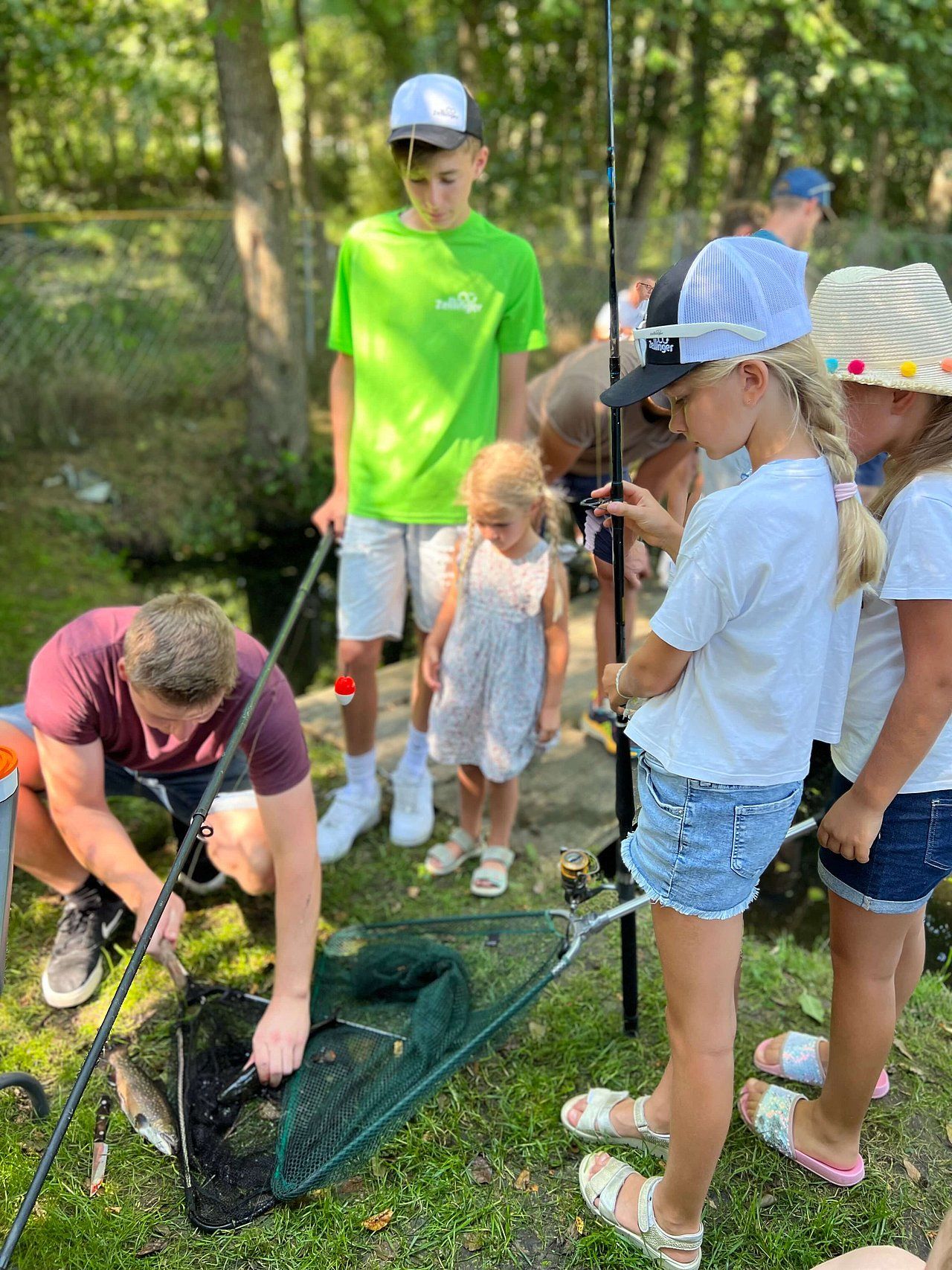 Group of children fishing with an adult. They are standing by a stream, looking at a fish in a net.