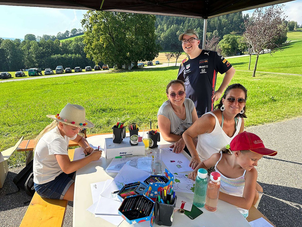 People at a table, coloring outdoors. Adults and children smile, surrounded by green fields and trees.