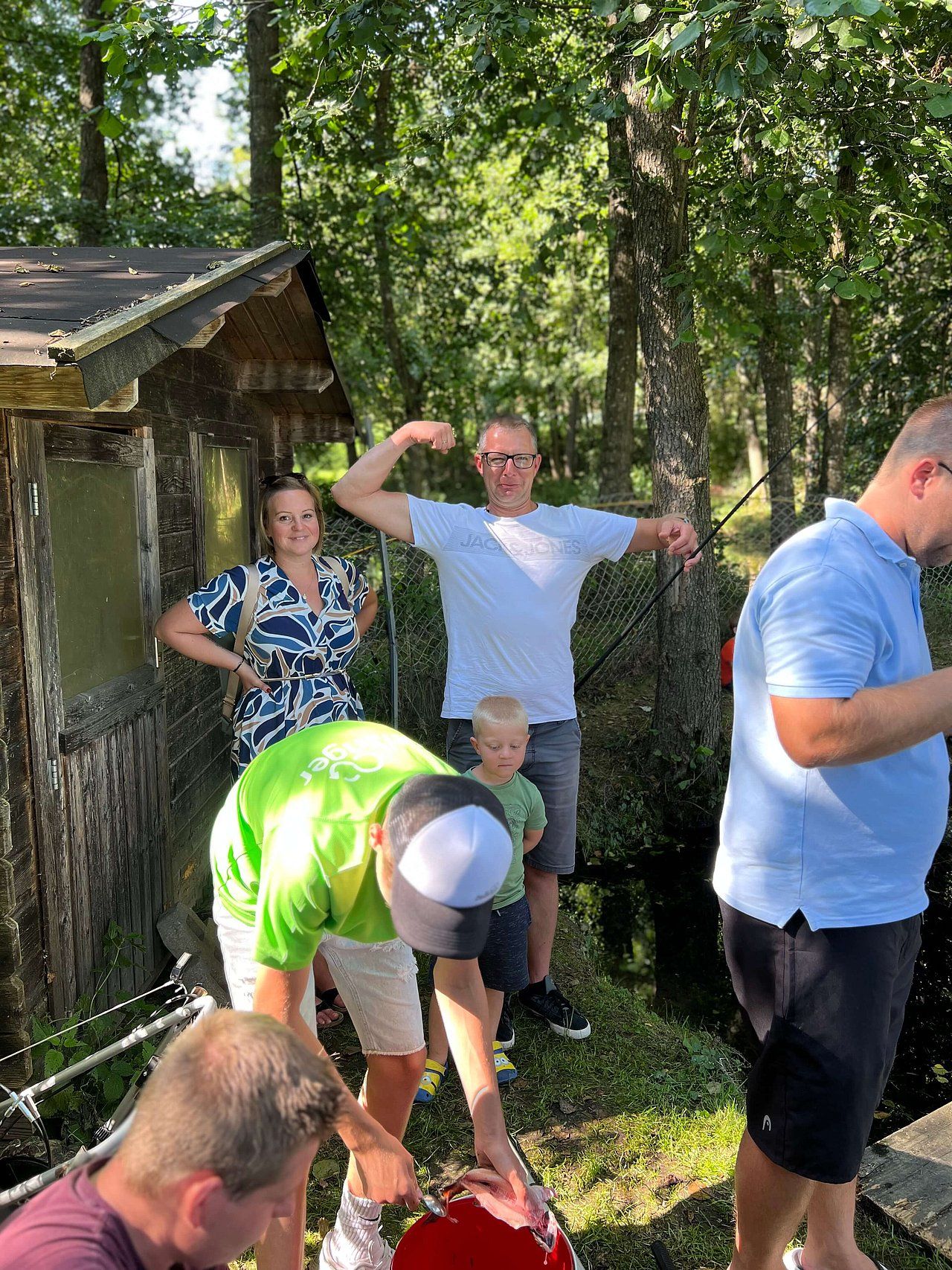 People gathered outdoors. Man flexes muscles; others stand and bend. A building and trees in the background.