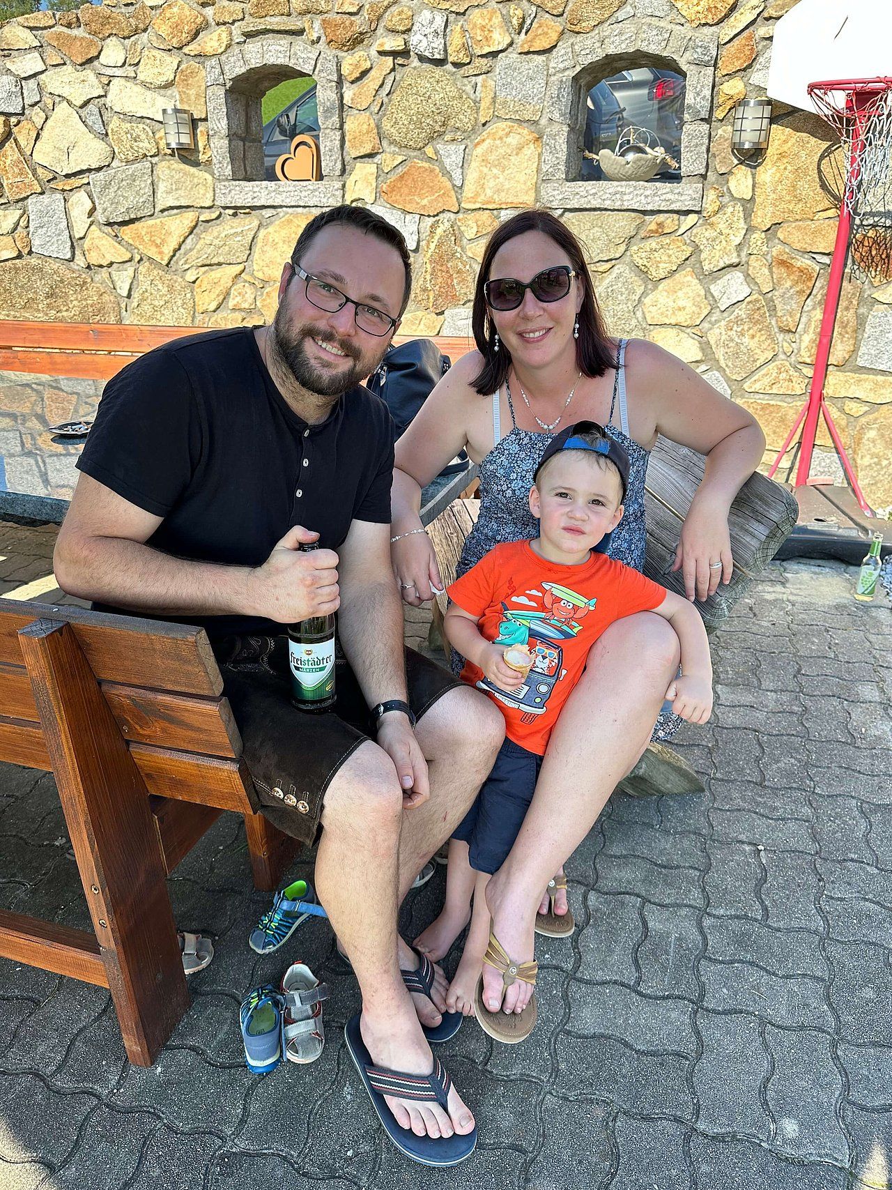 Family of three sitting outdoors: Man holding beer, woman smiling, child in orange shirt.