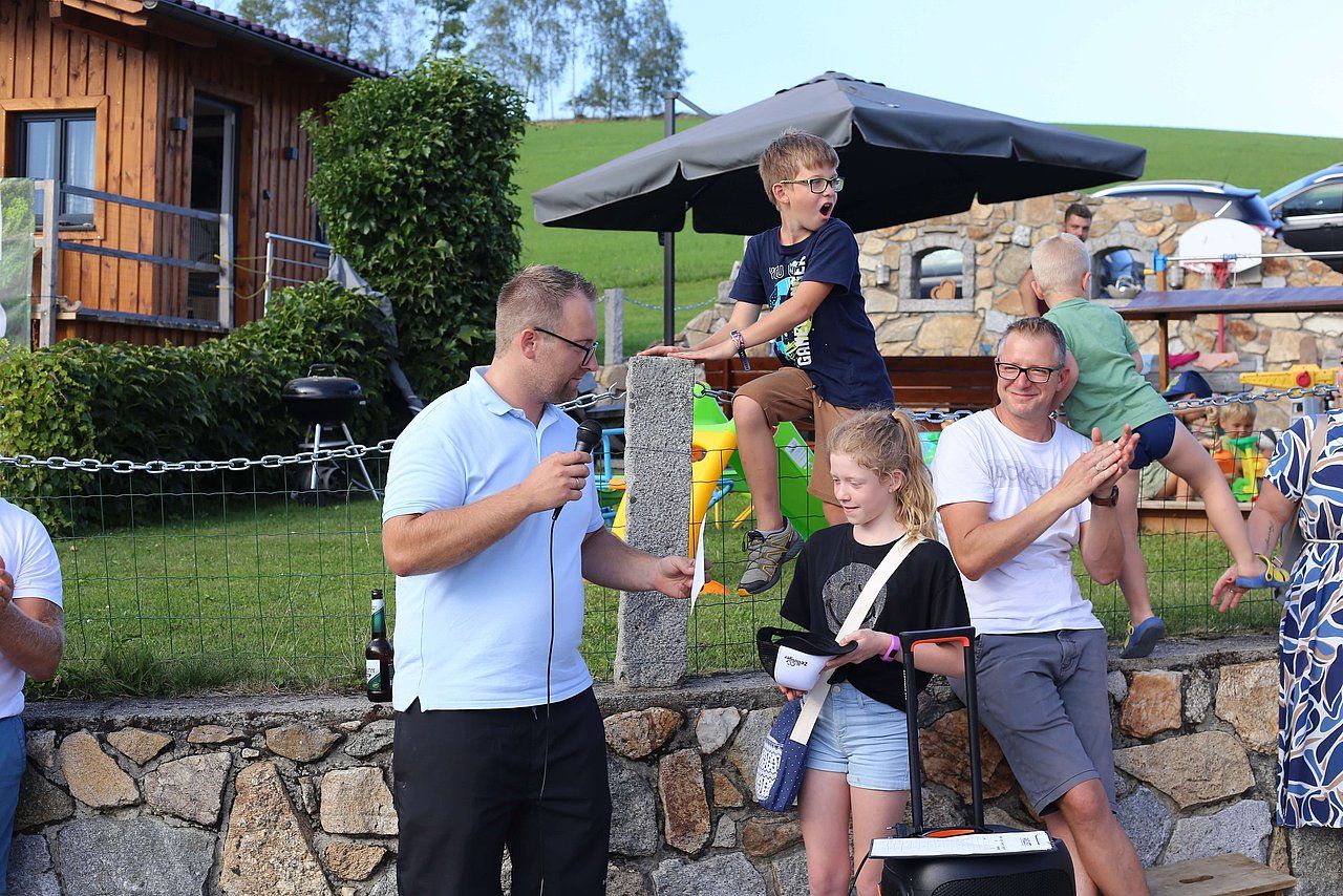Man with microphone speaks at an outdoor event; people listen, kids play nearby.