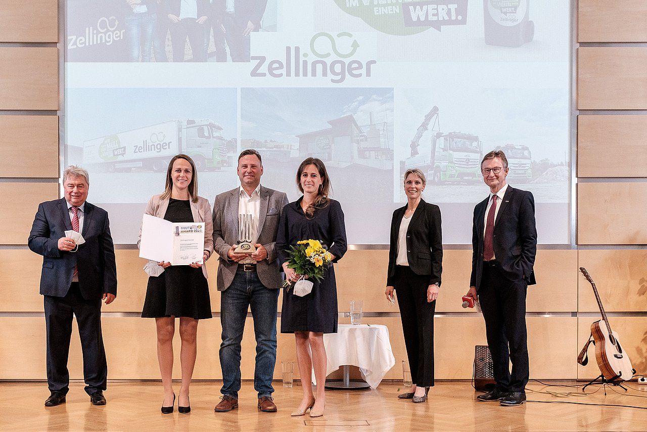 People on a stage receiving an award. A man and woman are in the center, holding a trophy and flowers.